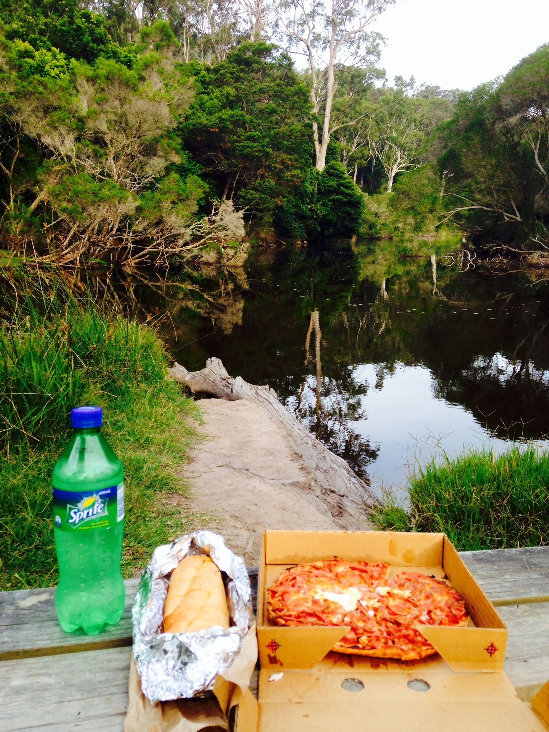 A picnic setup by a river with a Sprite bottle, a wrapped sandwich, and a pizza box.