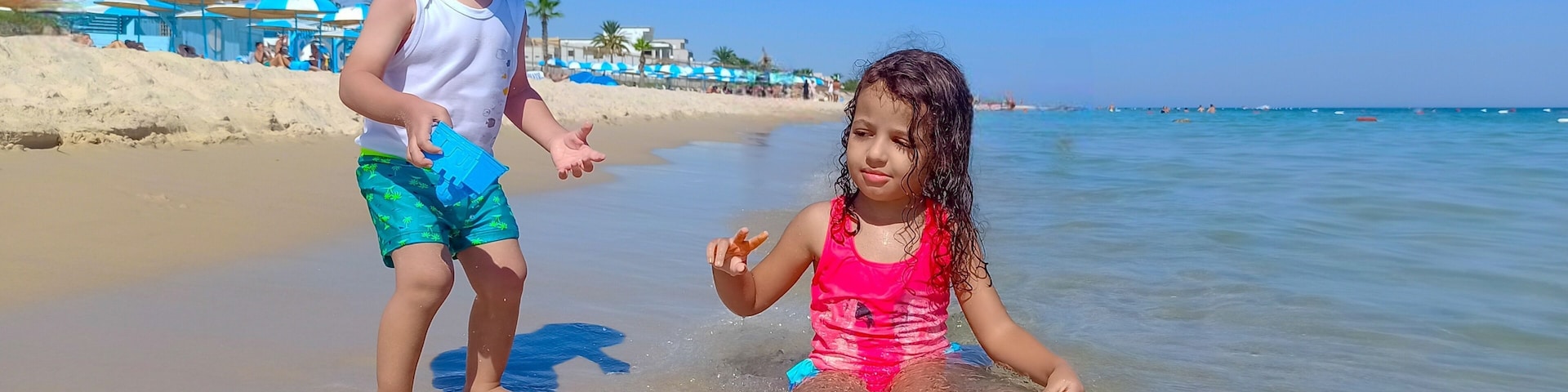 Two children playing in shallow water at a beach with umbrellas and palm trees in the background.