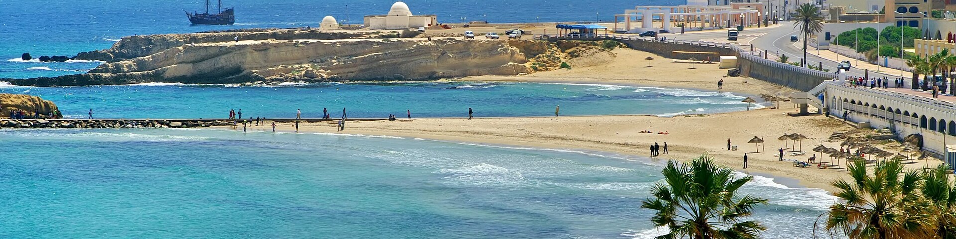 Un paisaje urbano costero con playa, palmeras y un barco a lo lejos.