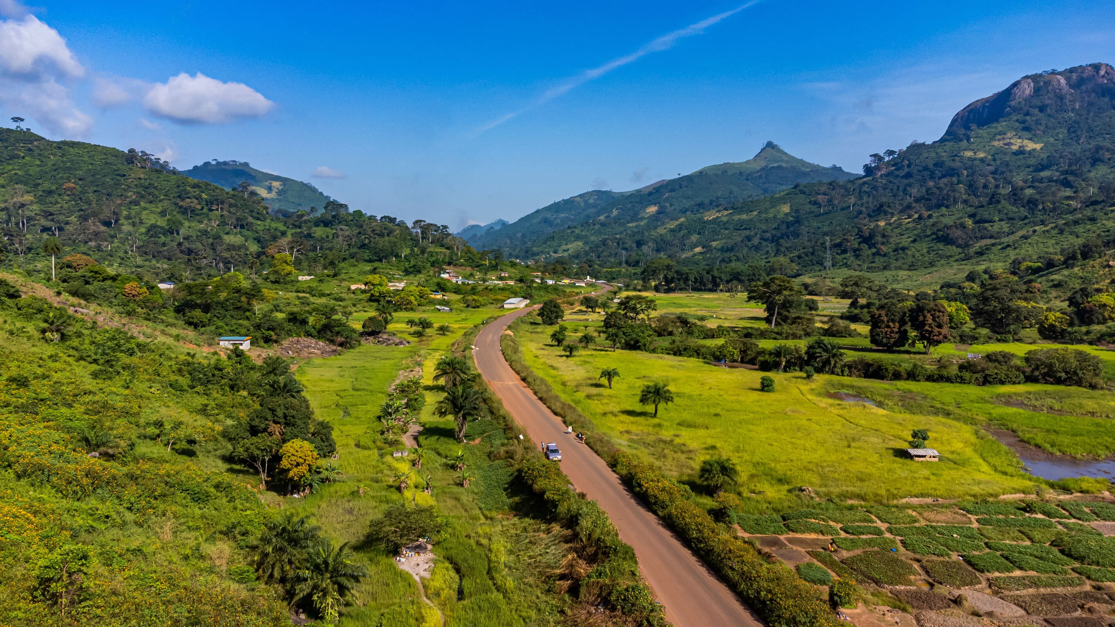 A winding road through a lush valley with green fields, scattered trees, and distant mountains under a clear blue sky.