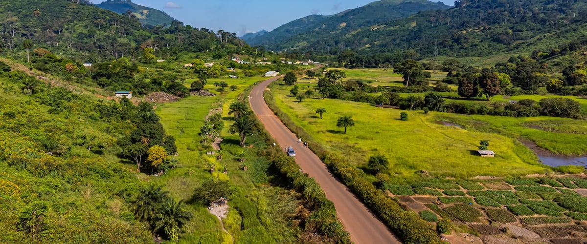 A winding road through a lush valley with green fields, scattered trees, and distant mountains under a clear blue sky.