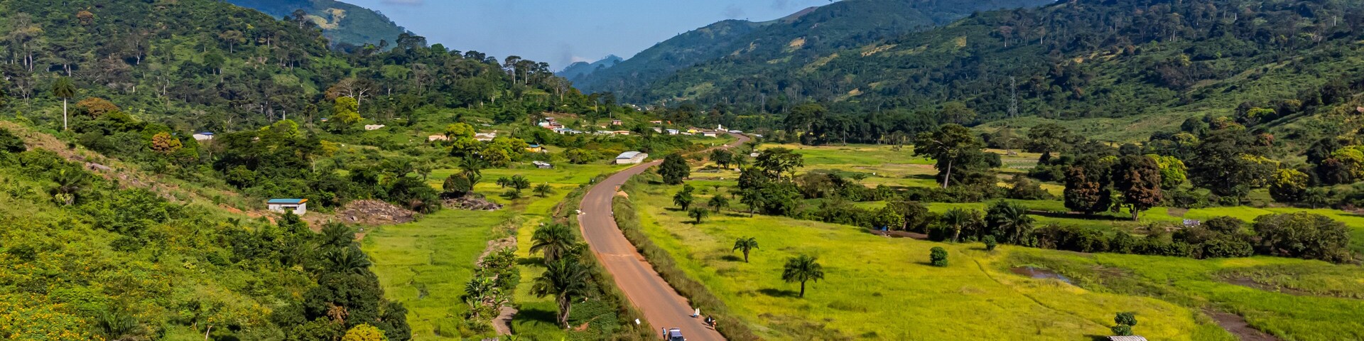 A winding road through a lush valley with green fields, scattered trees, and distant mountains under a clear blue sky.