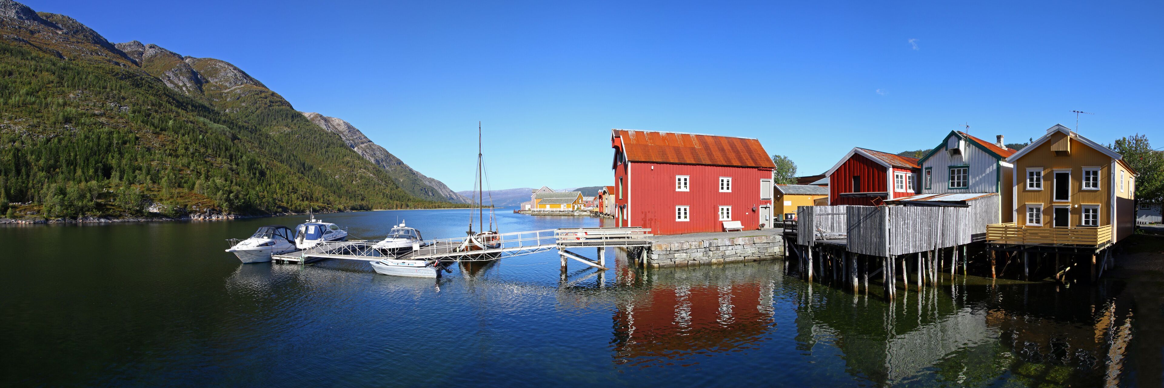 A calm lake with boats docked at a pier, surrounded by colorful houses and mountains.