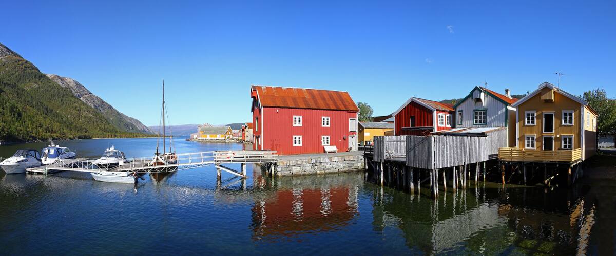 A calm lake with boats docked at a pier, surrounded by colorful houses and mountains.