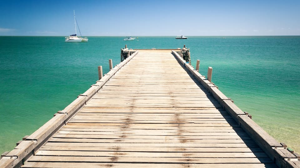 A wooden pier extending into turquoise water with sailboats in the distance.
