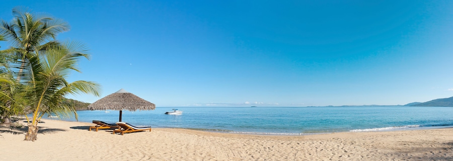 Uma praia com cadeiras de praia, um guarda-sol de palha, uma palmeira e um barco na água.