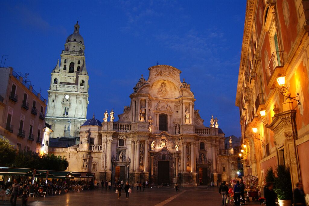 A historic cathedral with a bell tower and ornate facade, surrounded by a square with people and cafes.