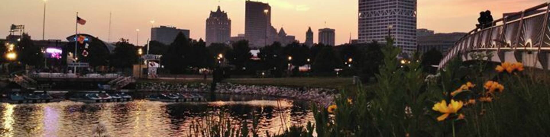 A city skyline at sunset with a river in the foreground and a bridge in the middle ground.