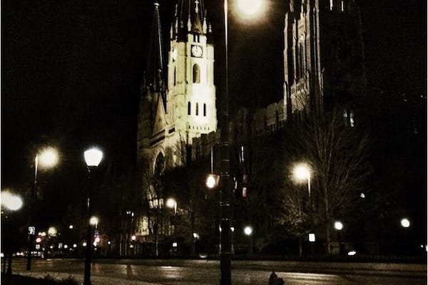 A cathedral at night with a large clock tower, surrounded by street lamps and a wet pavement.