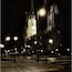 Uma catedral à noite com uma grande torre do relógio, cercada por luminárias de rua e calçada molhada.