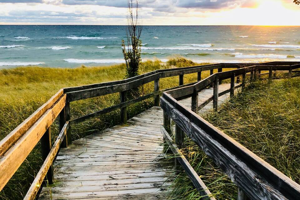 A wooden boardwalk leading to the ocean with grassy banks and a cloudy sky.