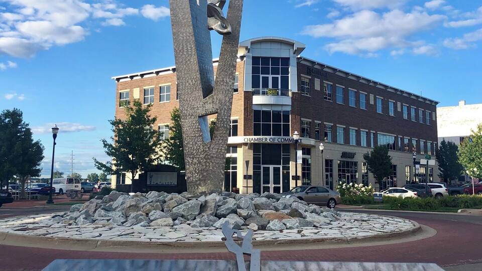 A sculpture titled 'MUSKEGON, TOGETHER RISING' by Richard Hunt, located in front of a building with 'CHAMBER OF COMMERCE' signage.