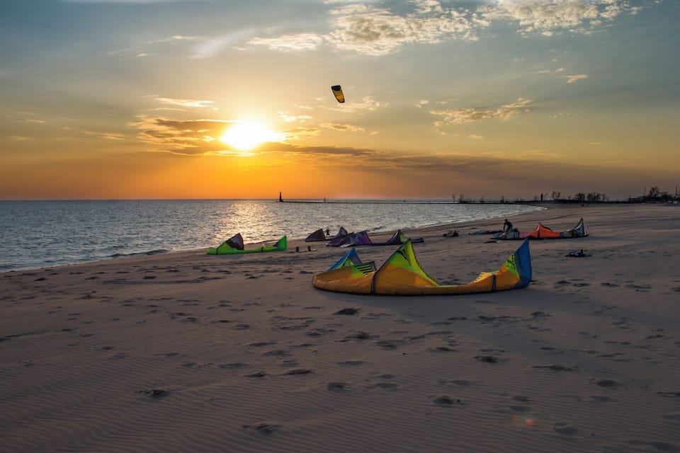 A beach at sunset with colorful kites and a silhouette of a lighthouse in the distance.