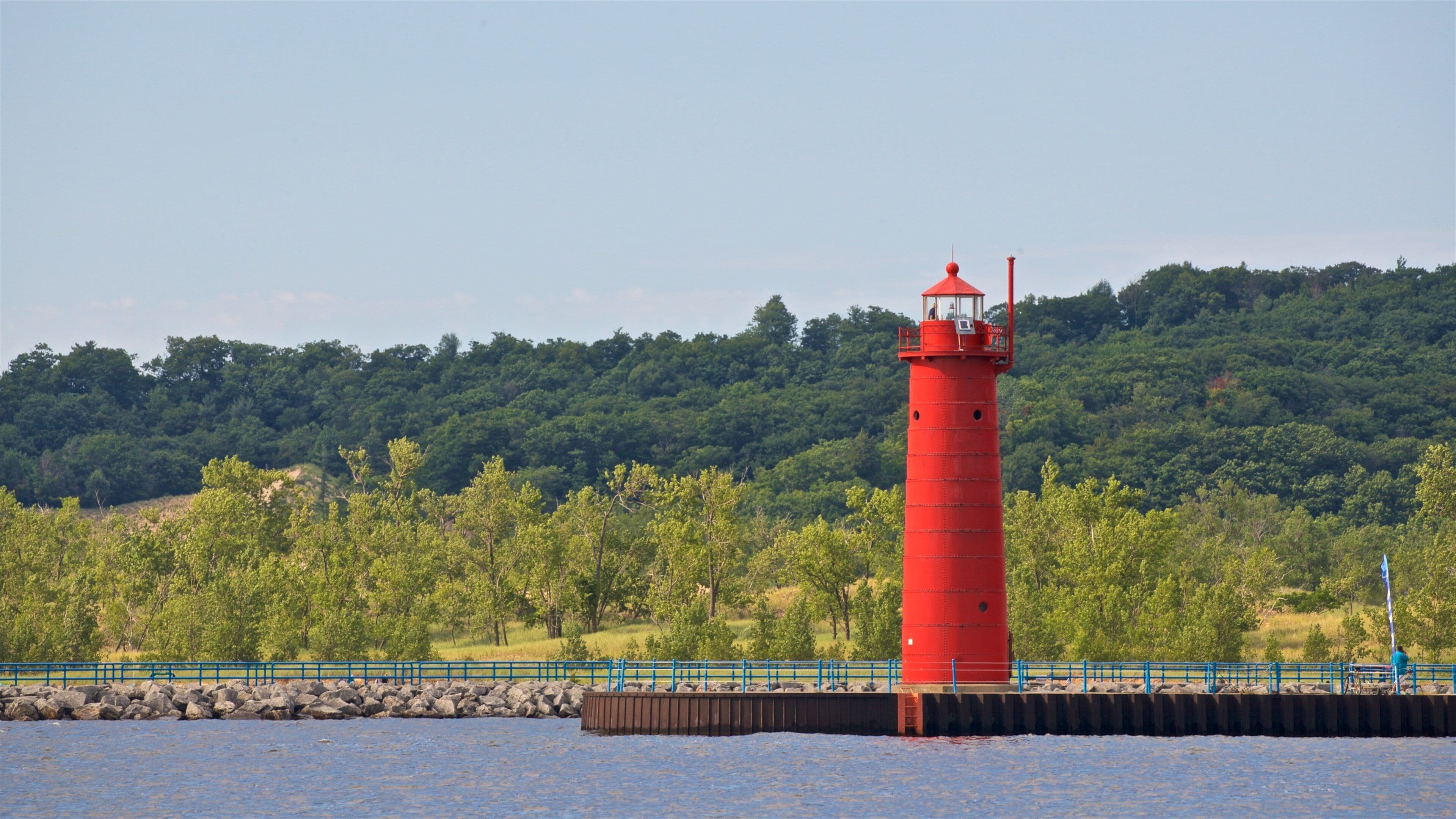 A red lighthouse by a calm body of water, surrounded by lush greenery and a forested hill.