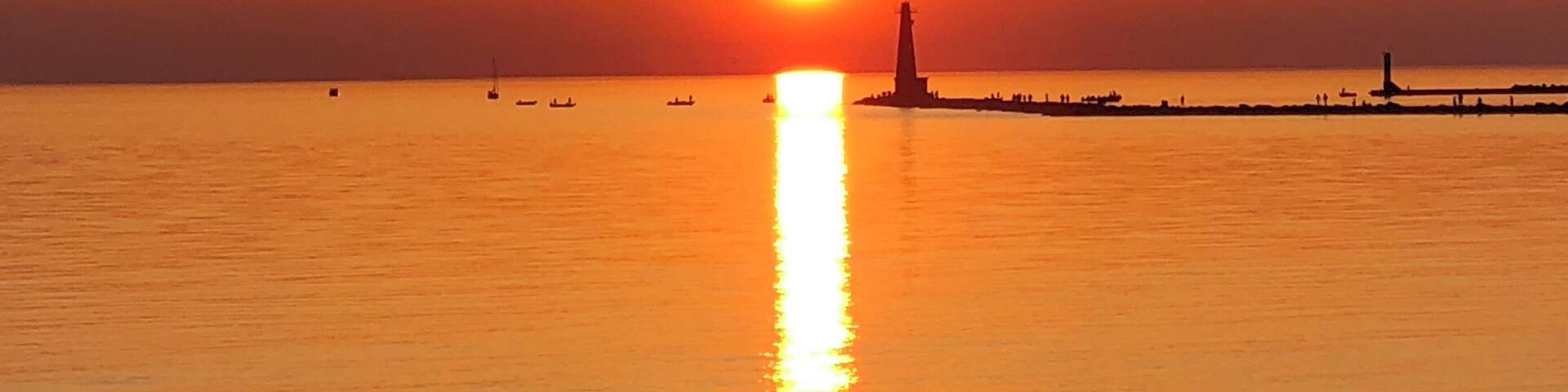 A sunset over a calm sea with a lighthouse and several boats.