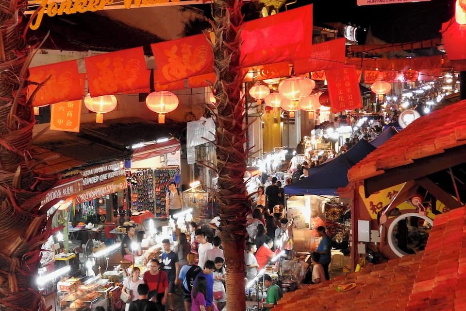 A bustling night market with red lanterns and signs, crowded with people and vendors.