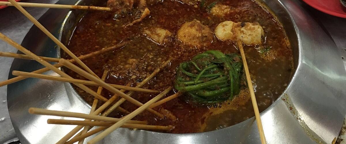 A metal pot with a stew containing meat and vegetables, surrounded by skewers and a knife.