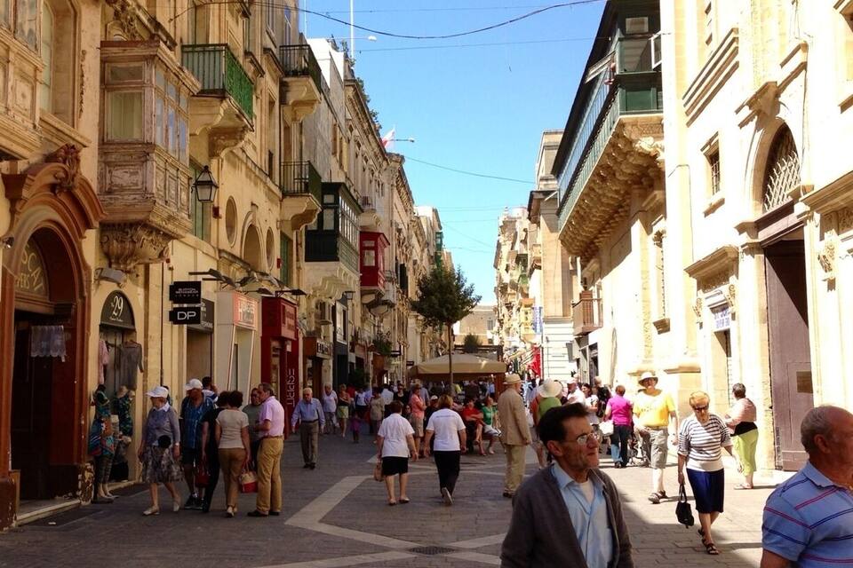 Rua movimentada com edifícios históricos, céu azul claro e pedestres a caminhar.