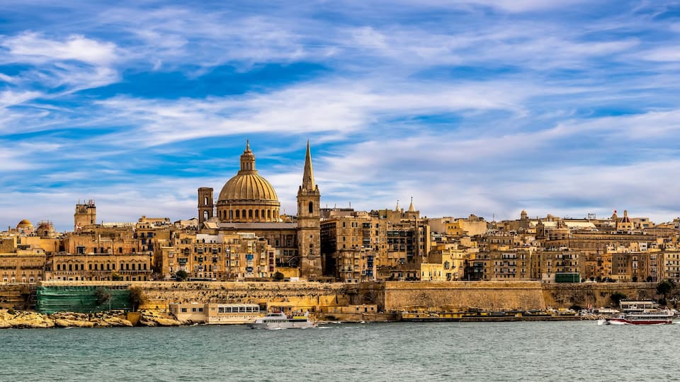A coastal cityscape with historic architecture, including a prominent dome and spires, under a partly cloudy sky.