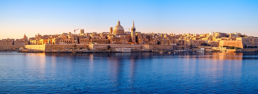 Paisagem costeira urbana com uma cúpula e torres imponentes, uma muralha fortificada e um mar calmo que reflete os edifícios.