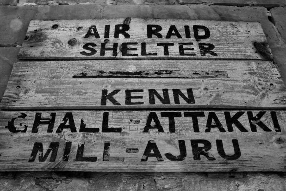 Ein hölzernes Schild mit dem Text 'AIR RAID SHELTER KENN CHALL-ATTAKKI MILI-AJRU'.