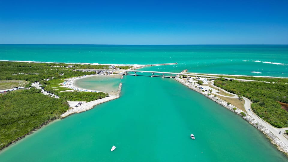 A coastal area with a bridge, turquoise waters, and a beach with parked cars.