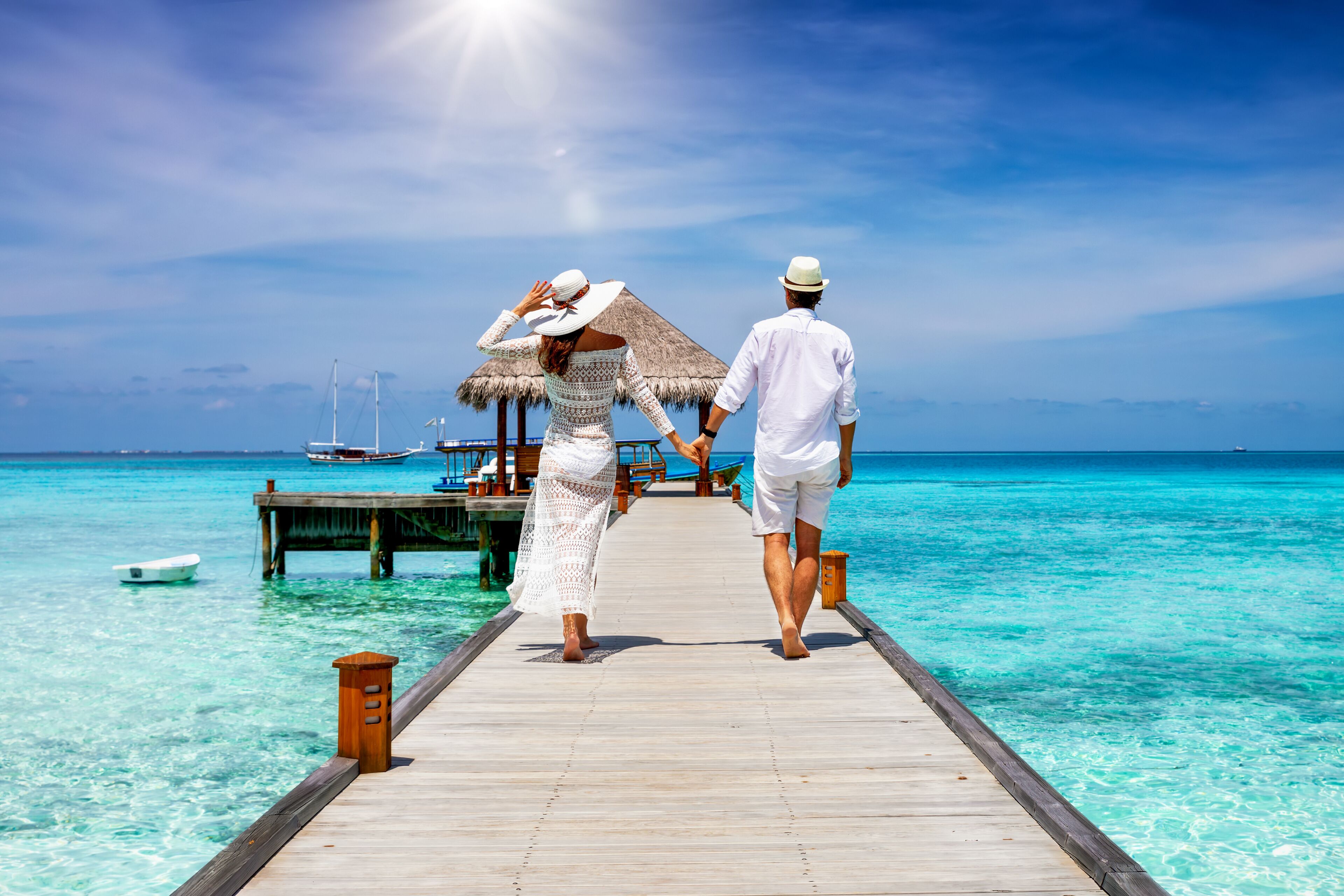 Una pareja caminando sobre un muelle de madera hacia una cabaña con techo de paja junto al mar turquesa.