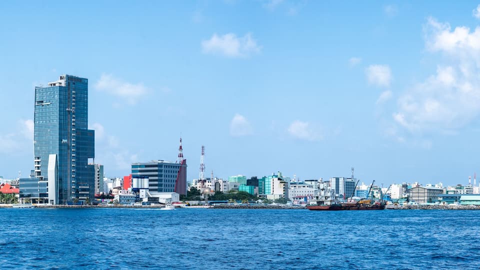 A coastal cityscape with a prominent skyscraper, a harbor with ships, and a clear blue sky.