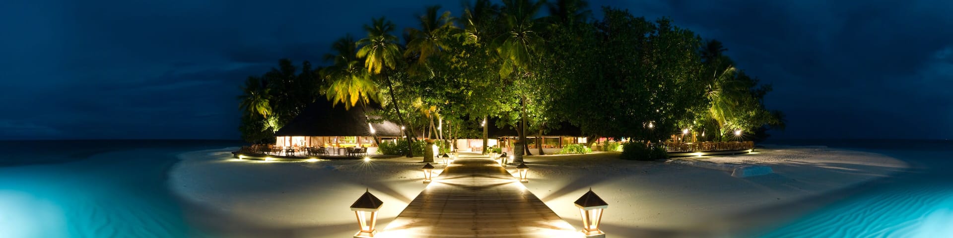A wooden pathway leading to a beachside resort with palm trees and thatched-roof structures.