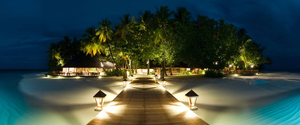A wooden pathway leading to a beachside resort with palm trees and thatched-roof structures.
