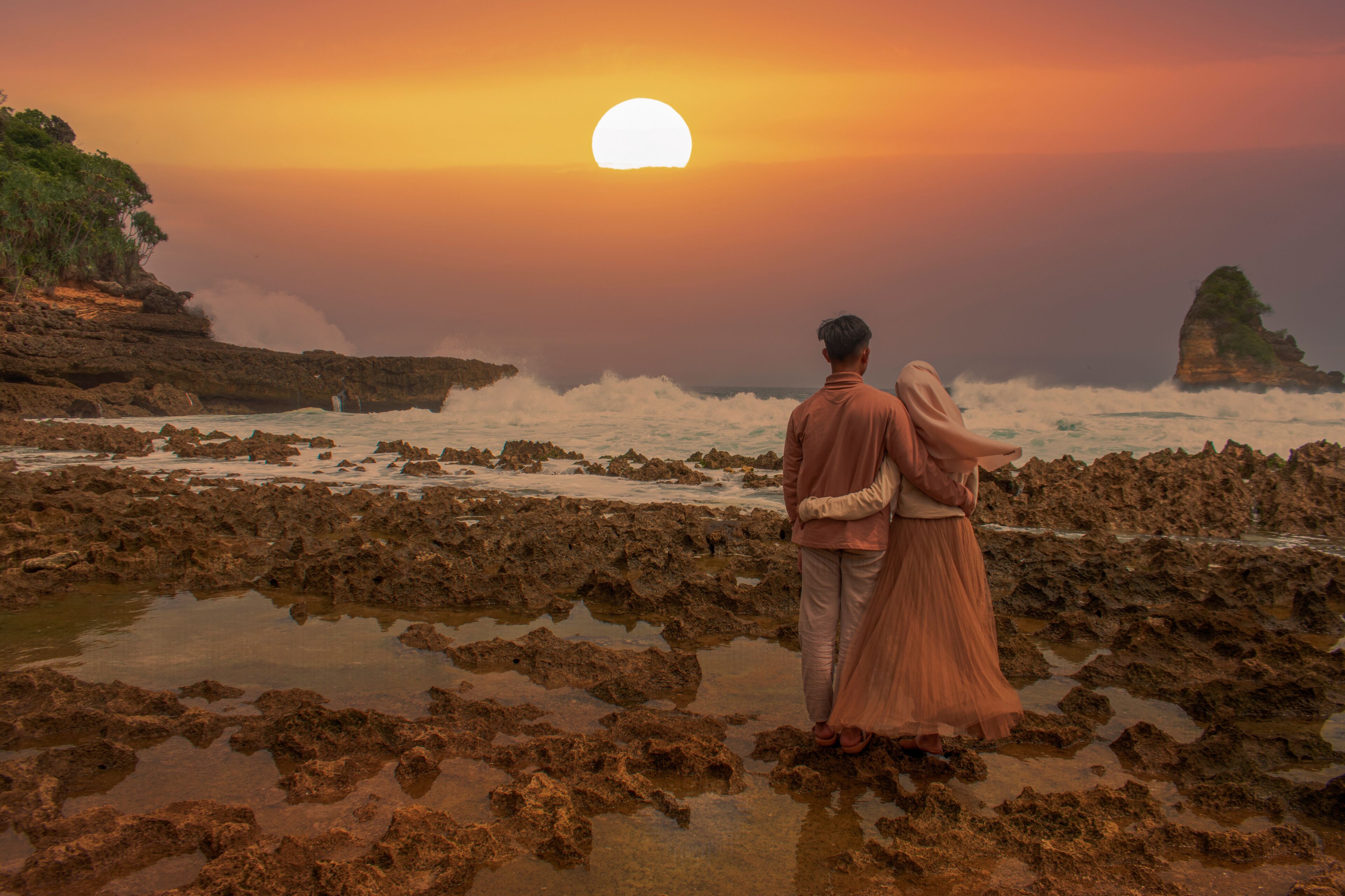 A couple standing on rocky terrain with waves crashing nearby, under a sunset sky.