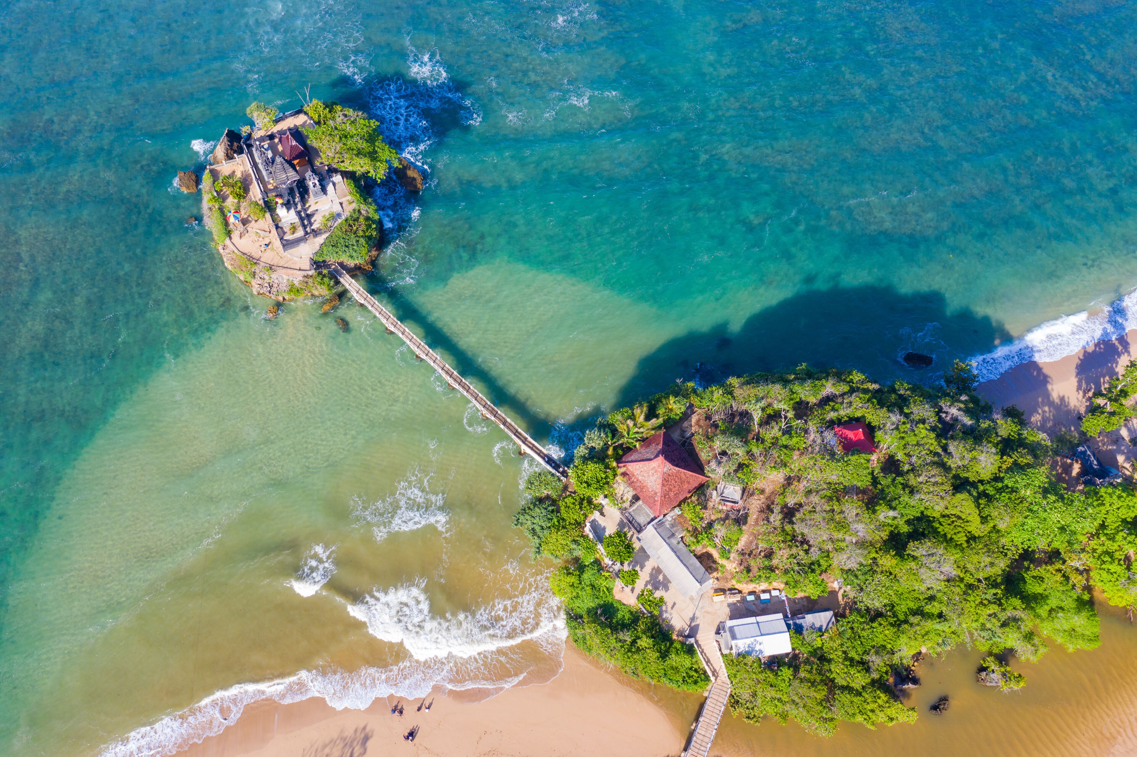 An aerial view of a coastal area with a beach, a pier, and a small island with buildings.
