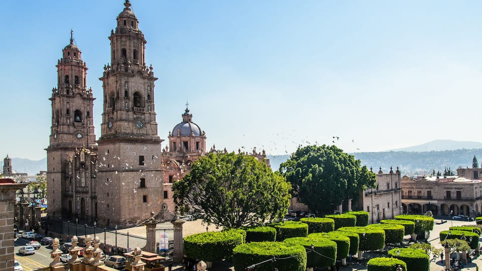 A historic cathedral with two prominent towers and a large dome, surrounded by a well-maintained square with trimmed hedges.