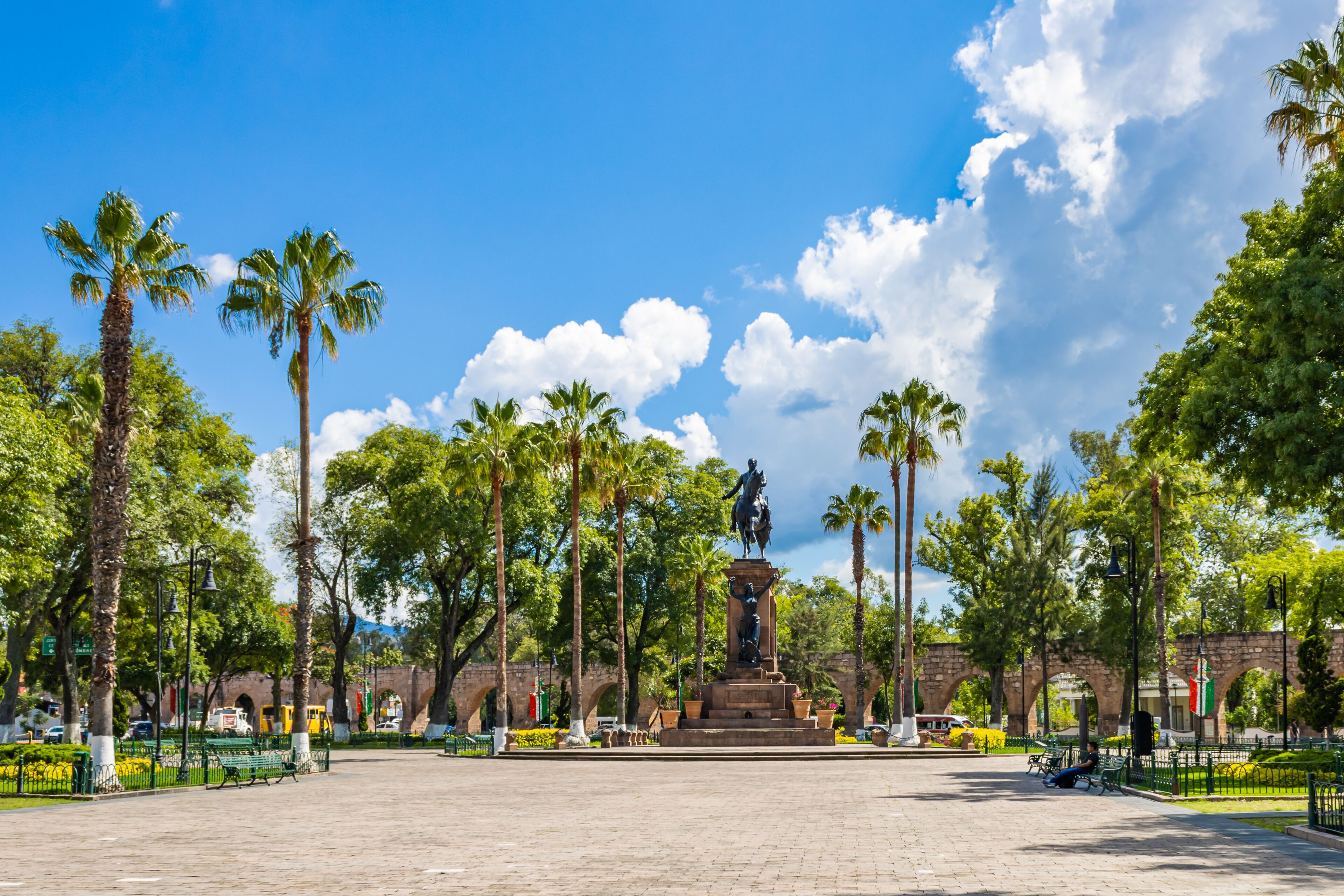 Uma praça com uma estátua central, palmeiras e um céu azul límpido.