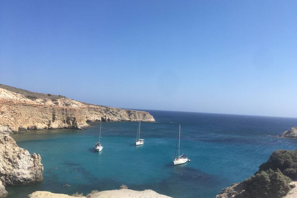 Un paisaje costero con aguas azules cristalinas, acantilados rocosos y tres veleros anclados cerca de la orilla.