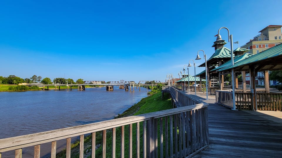 A wooden boardwalk along a river with a bridge in the background.