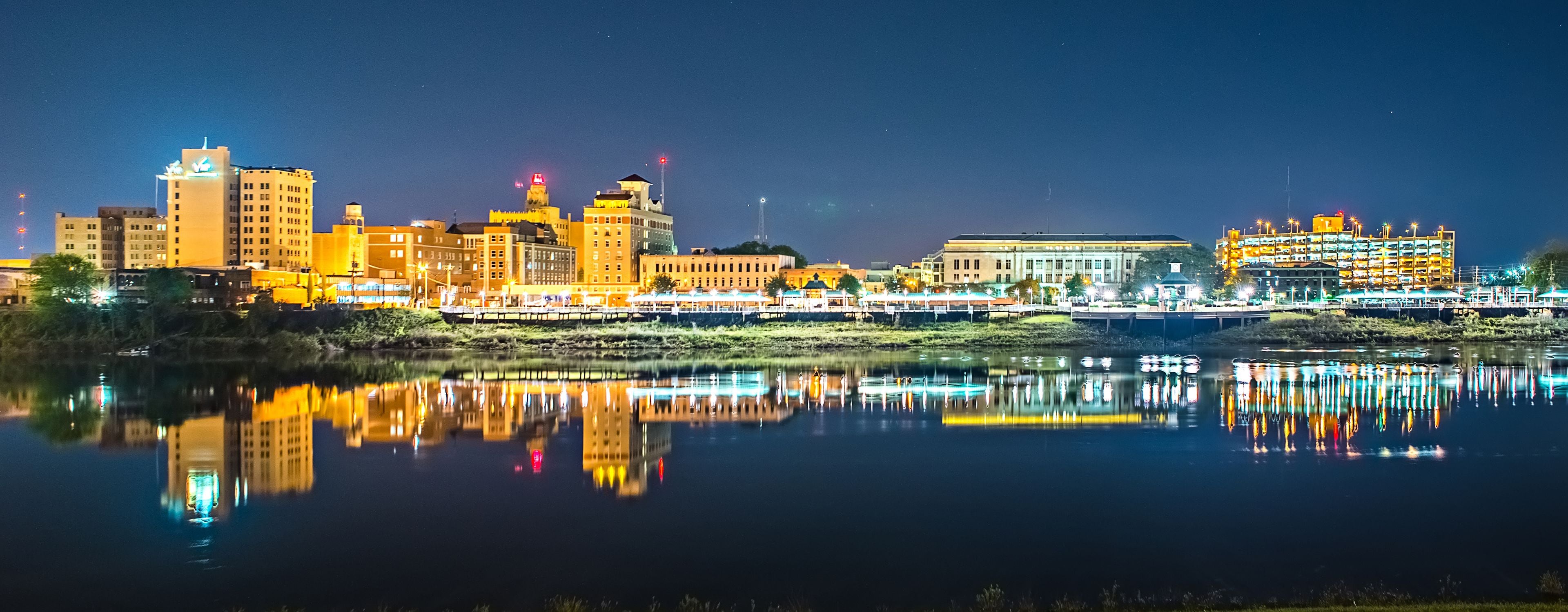 A city skyline at night with buildings reflected in a calm body of water.