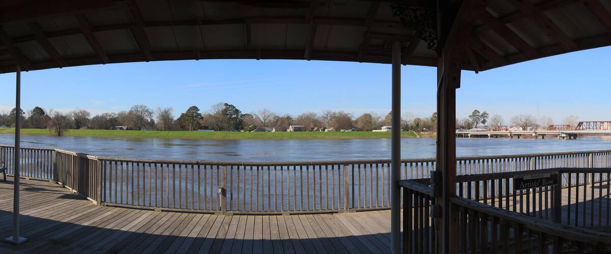 A wooden deck overlooking a river with a metal railing, a grassy area, and a bridge in the background.