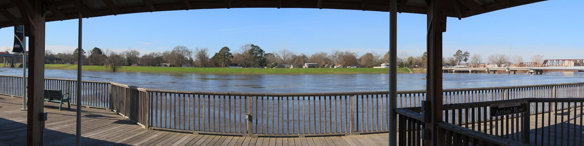A wooden deck overlooking a river with a metal railing, a grassy area, and a bridge in the background.
