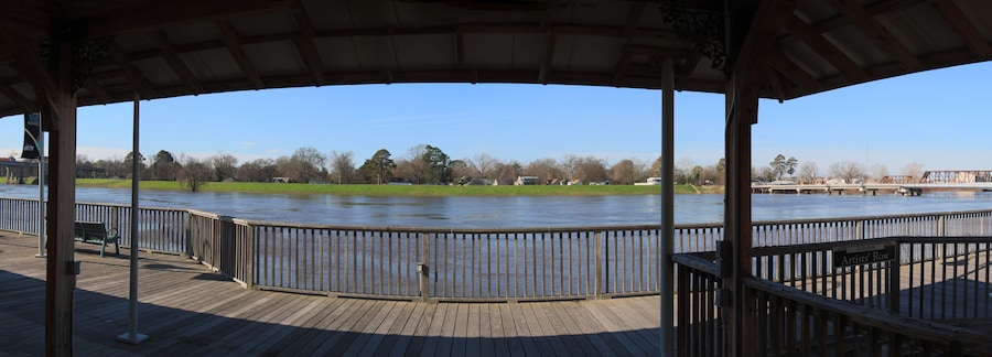 Una terraza de madera con vistas a un río, con barandilla de metal, zona verde y un puente al fondo.