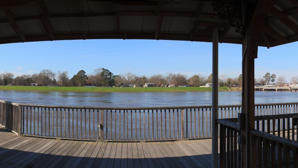 A wooden deck overlooking a river with a metal railing, a grassy area, and a bridge in the background.