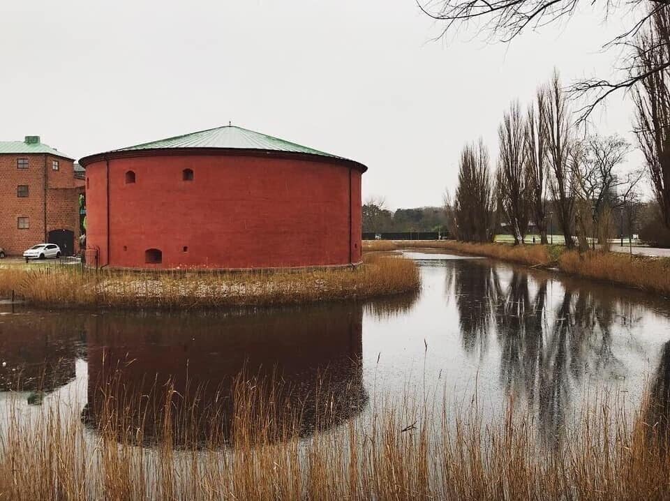 A red cylindrical building near a body of water with reflections, surrounded by bare trees and a brick building in the background.
