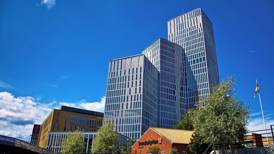 Un edifici modern de vidre amb un disseny angular únic, un edifici de maó tradicional amb un rètol, i un cel blau clar.