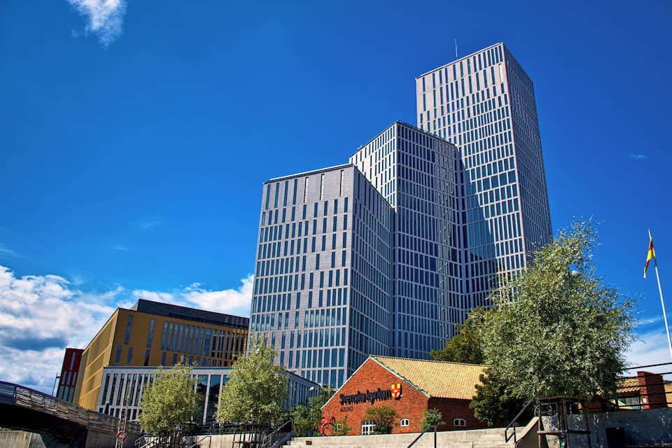 A modern glass building with a unique angular design, a traditional brick building with a sign, and a clear blue sky.