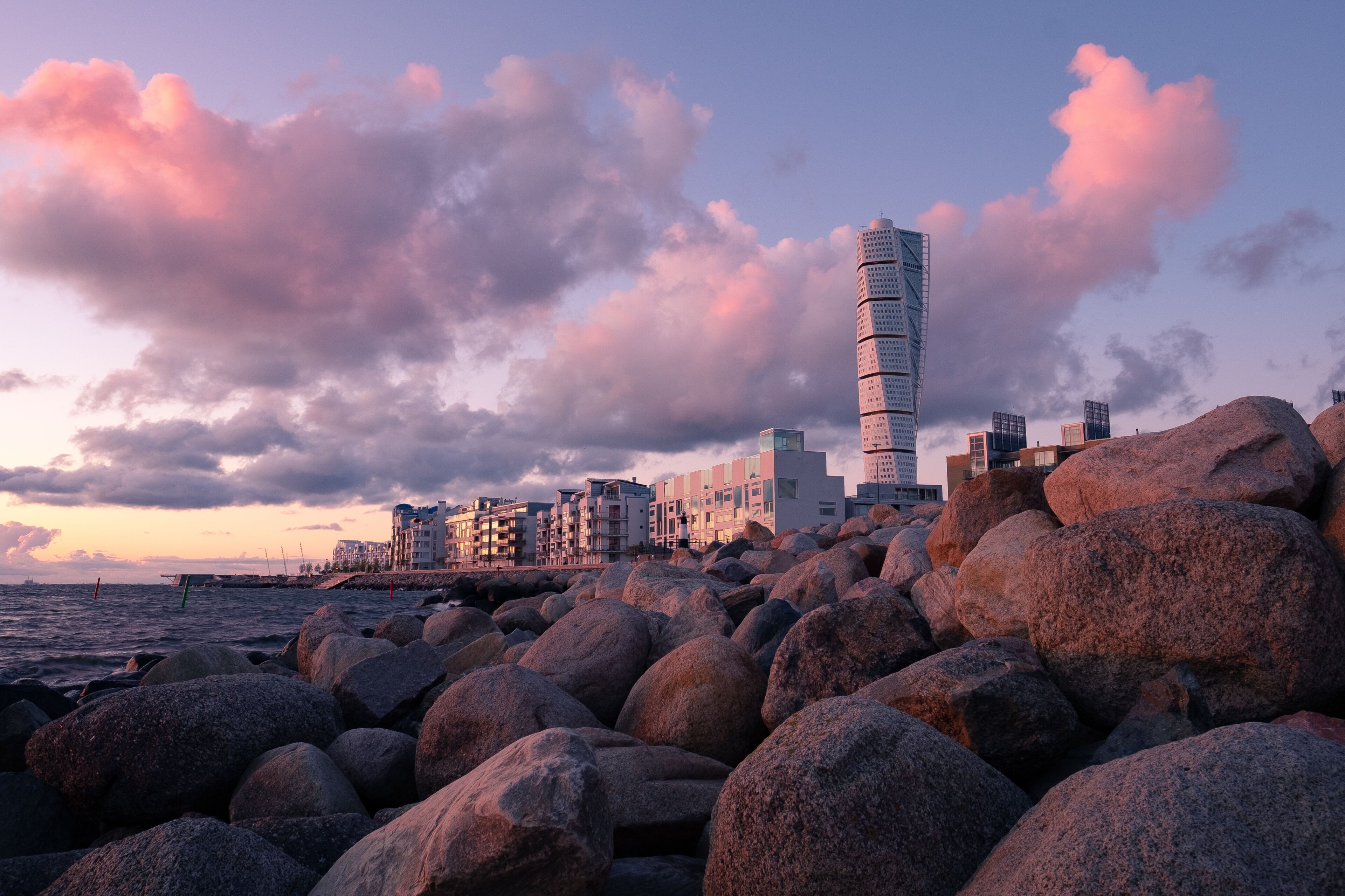 Un paisaje urbano costero con una torre retorcida destacada, rocas grandes y edificios modernos bajo un cielo de tonos rosados.
