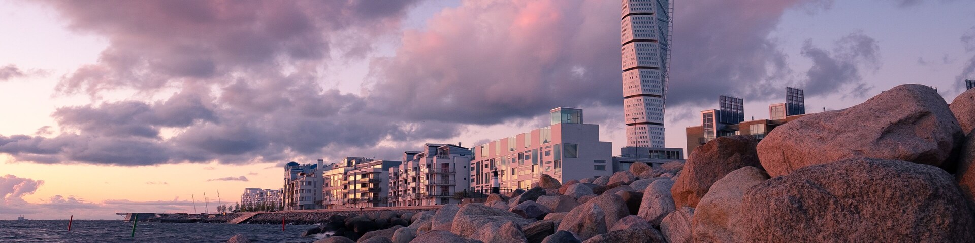 Un paisaje urbano costero con una torre retorcida destacada, rocas grandes y edificios modernos bajo un cielo de tonos rosados.
