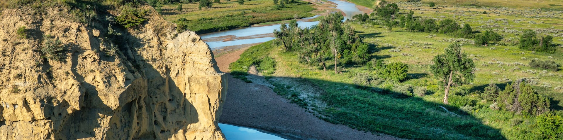 A river flowing through a valley with a cliff on the left and a grassy landscape with trees.