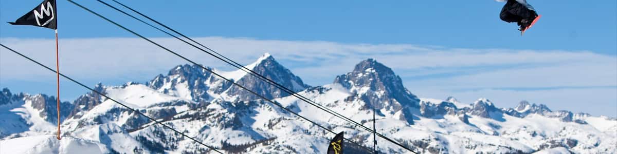 Un snowboarder en plein saut, avec en arrière-plan une montagne enneigée, un télésiège et des drapeaux.