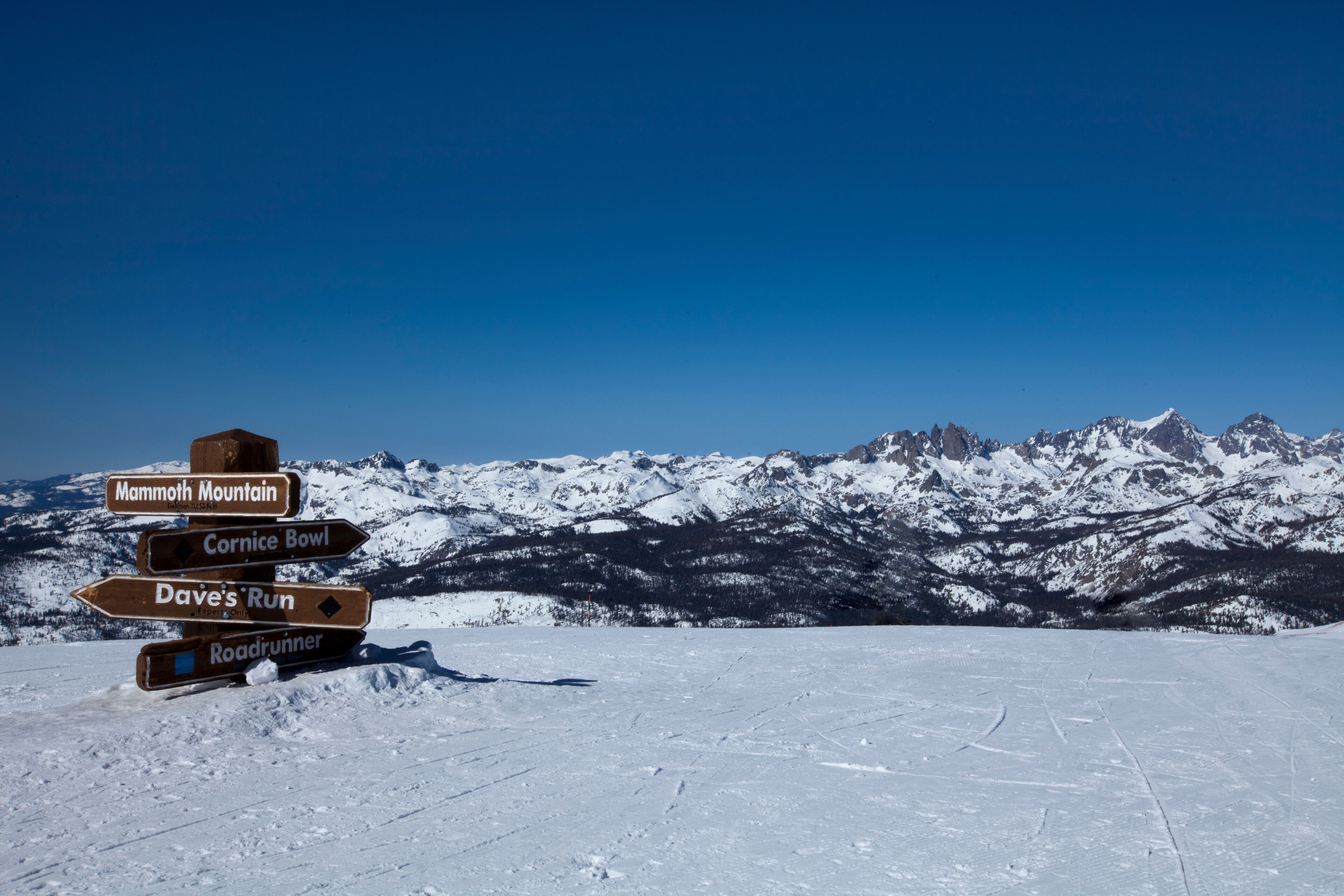 Paisaje nevado de montaña con un poste de madera que indica direcciones a Mammoth Mountain, Cornice Bowl, Dave's Run y Roadrunner.