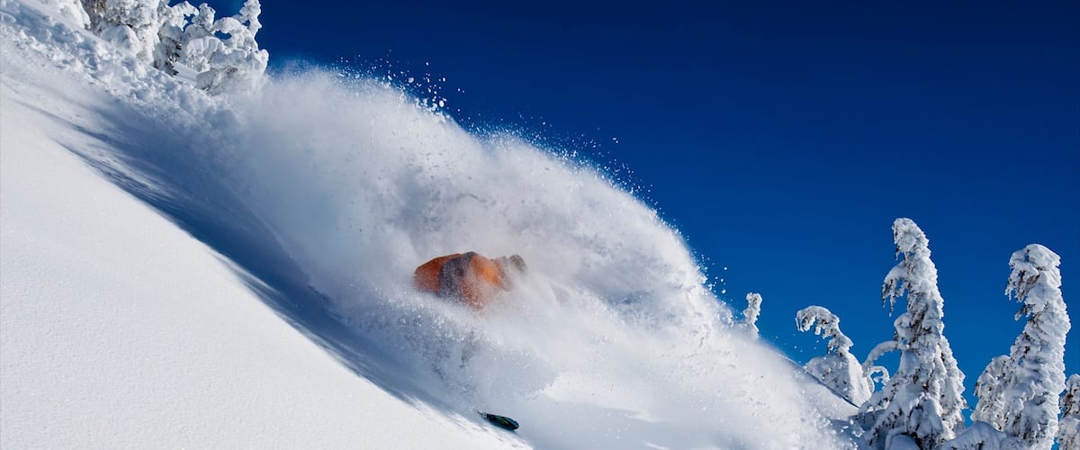 A snowboarder carving through deep snow with a clear blue sky in the background.